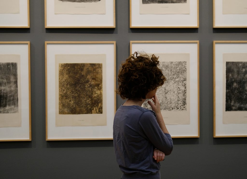 Persona de cabello rizado observa obras sobre papel enmarcadas en una pared gris.