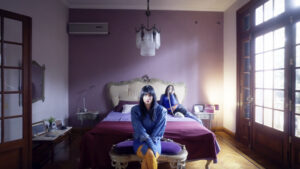 Florencia Hana Ciliberti sentada en un banco al pie de una cama, vestida de azul, con otra mujer al fondo en una habitación de paredes lilas.