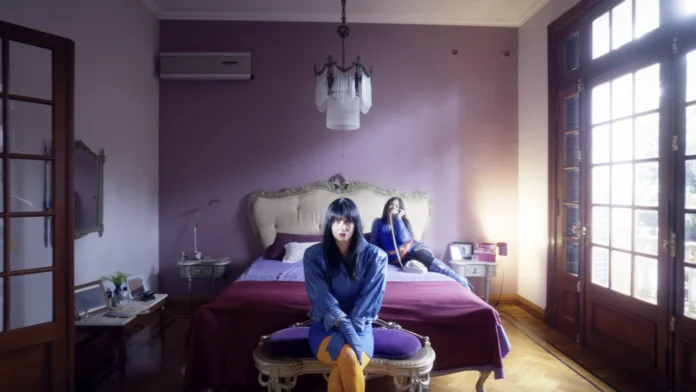Florencia Hana Ciliberti sentada en un banco al pie de una cama, vestida de azul, con otra mujer al fondo en una habitación de paredes lilas.
