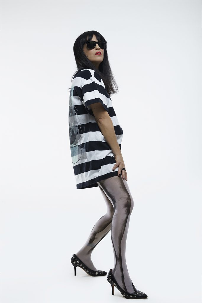 Retrato de cuerpo entero de Hana Ciliberti con lentes oscuros, camiseta de rayas blancas y negras, medias brillantes y zapatos con estrellas.