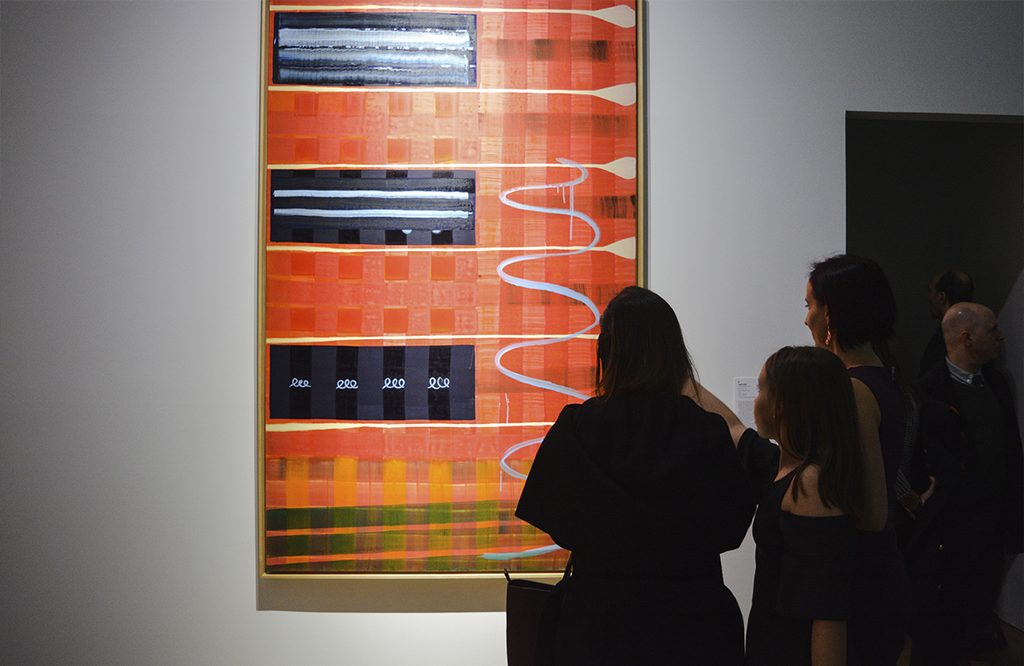 Visitantes observan una obra abstracta de gran formato con colores naranjas, líneas y formas geométricas.