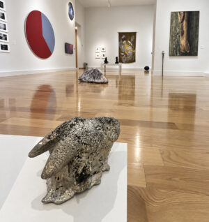 Escultura de apariencia metálica sobre un pedestal blanco, con obras en las paredes y el piso de una sala de exposición.