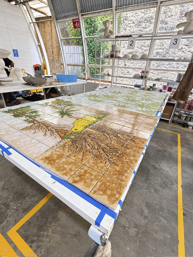 Panel de piezas cuadradas con una composición en tonos verdes, amarillos y cafés sobre una mesa de trabajo en Cerámica Suro.
