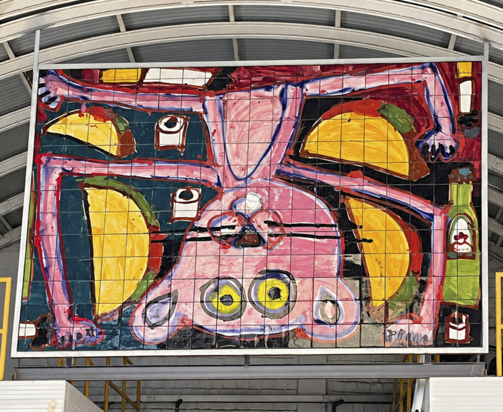 Mural formado por piezas cuadradas con figuras delineadas en negro y colores rosa, rojo, amarillo y verde, montado sobre una estructura metálica.