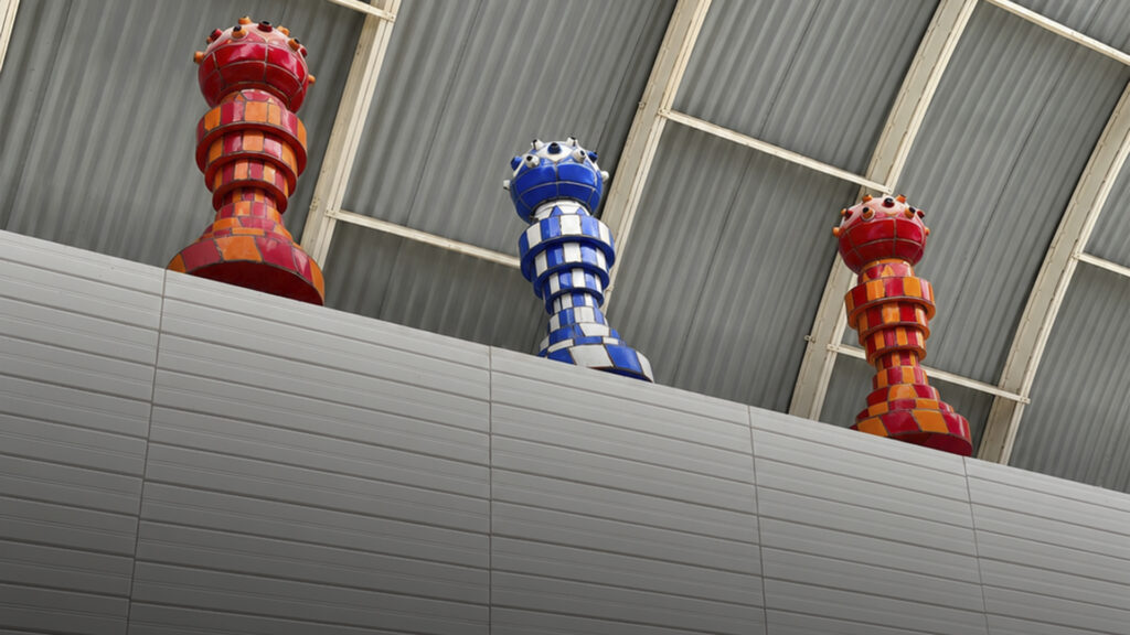 Tres piezas escultóricas verticales con patrones rojos, amarillos, azules y blancos, colocadas sobre una estructura elevada dentro de la fábrica.