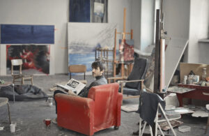 Persona sentada en un sillón rojo dentro de un taller con pinturas, caballetes y materiales de trabajo.