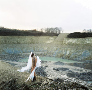 A figure seen from behind, with long hair and white, wing-like features, standing in front of a quarry filled with turquoise water.