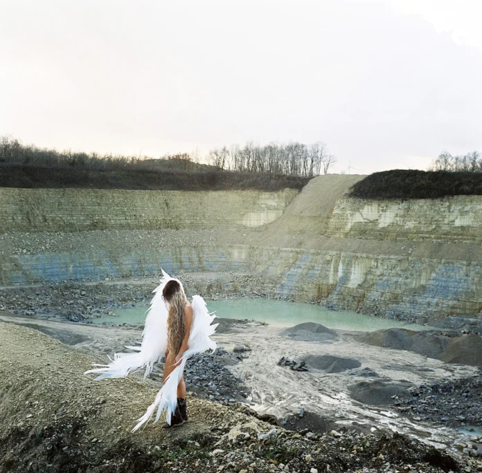 A figure seen from behind, with long hair and white, wing-like features, standing in front of a quarry filled with turquoise water.