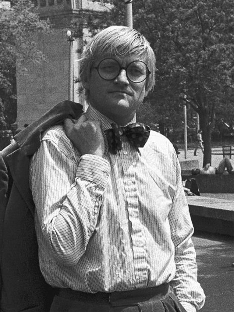 Retrato en blanco y negro de David Hockney con lentes redondos, camisa a rayas y moño, sosteniendo un saco sobre el hombro.