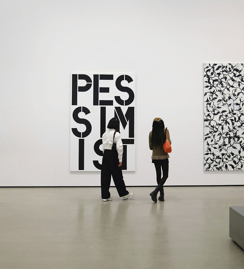 Dos visitantes contemplan obras de arte en una sala con paredes blancas y piso de madera.