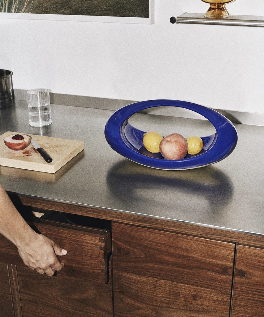 Frutero azul con cítricos y un durazno sobre una cubierta de cocina, junto a una mano que abre un cajón de madera.