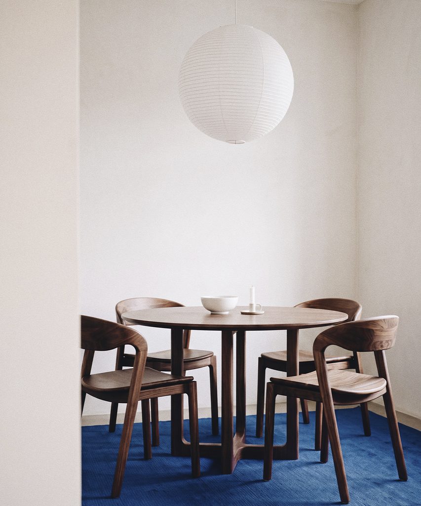 Sillas  de madera con asientos tejidos alrededor de una mesa baja cuadrada con un jarrón, en una sala con pintura abstracta.