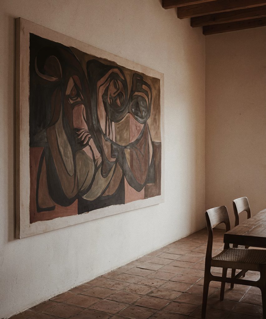 Dos sillones de madera con asientos tejidos alrededor de una mesa baja cuadrada con un jarrón, en una sala con pintura abstracta.