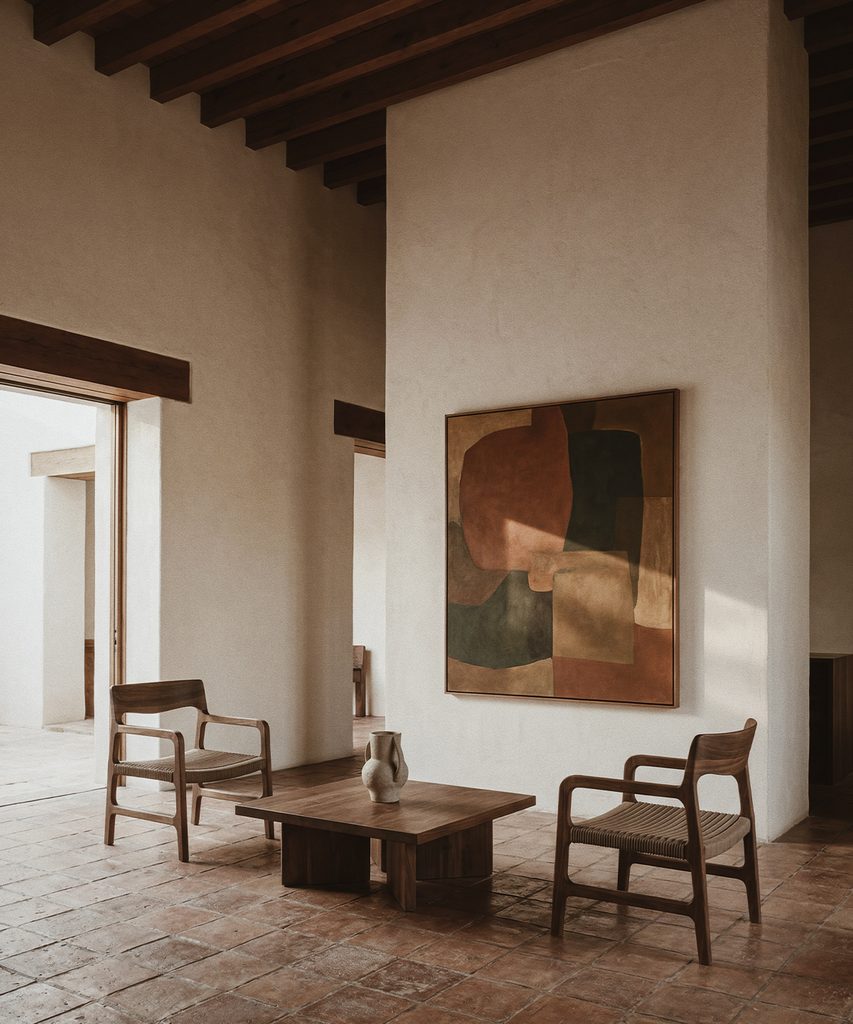 Sala con dos sillones de madera, mesa baja y jarrón, piso de terracota, vigas oscuras y una pintura abstracta.