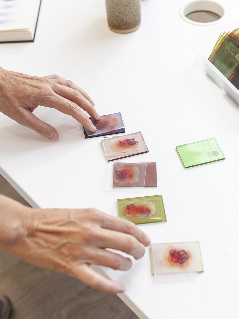 Hands arranging small rectangular pieces of glass with orange and violet spots on a white table.