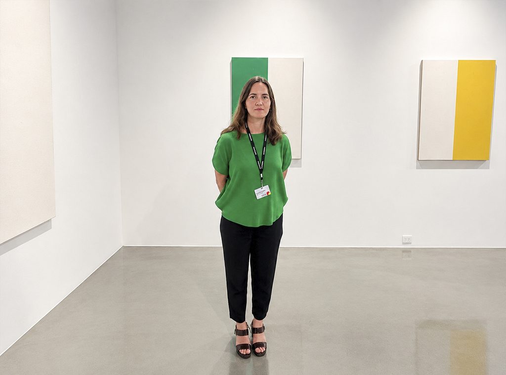 Claudia Llanza, wearing a green blouse and black pants, stands in front of paintings of white, green, and yellow fields.