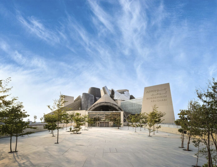 The facade of the Guggenheim Abu Dhabi, designed by Frank Gehry, featuring a glass entrance, sculptural forms, and a tree-lined plaza.