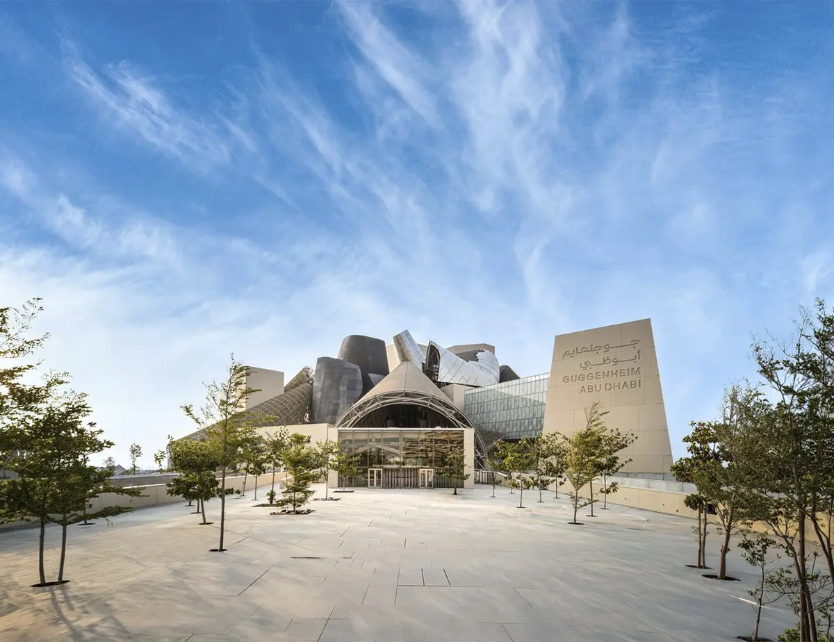 Fachada del Guggenheim Abu Dhabi diseñada por Frank Gehry, con entrada acristalada, volúmenes escultóricos y una plaza arbolada.