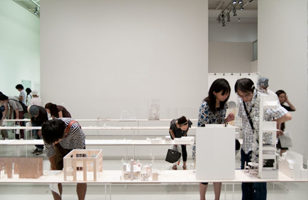 Visitors lean over white tables to examine architectural models in an exhibition hall.