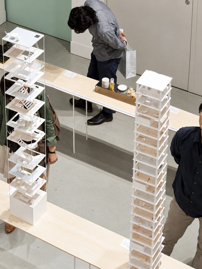 A bird's-eye view of two white models of tall buildings, with visible floors and staircases, surrounded by visitors at an exhibition.