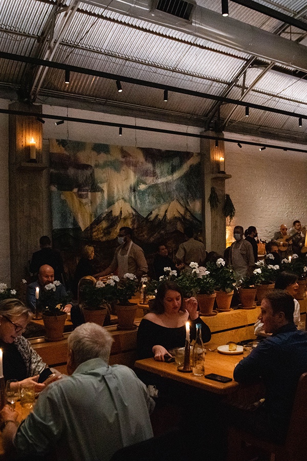 Comedor de Máximo con comensales, mesas de madera, cubierta alta y mural.