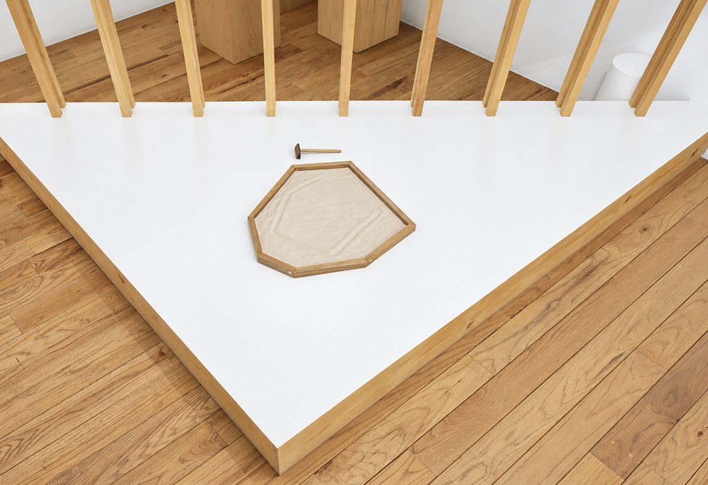 Pieza poligonal con marco de madera y superficie clara sobre una plataforma blanca, junto a una pequeña herramienta y una estructura de listones de madera.