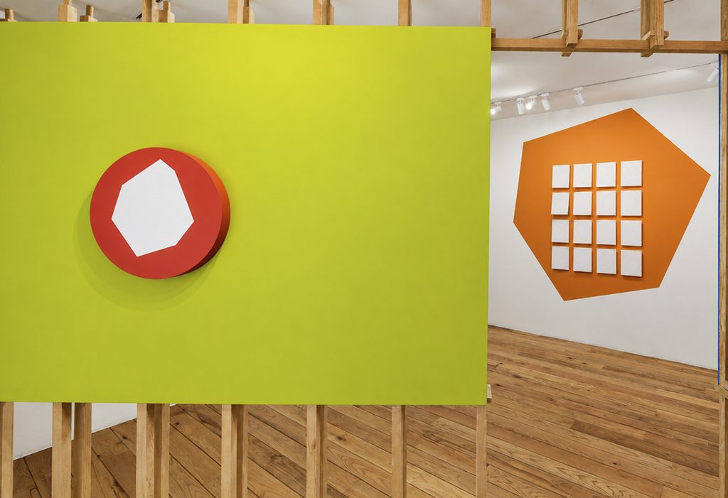 Pintura circular roja con un polígono claro sobre un panel verde amarillo; al fondo, dieciséis dibujos dispuestos en una cuadrícula sobre una figura naranja en el muro.