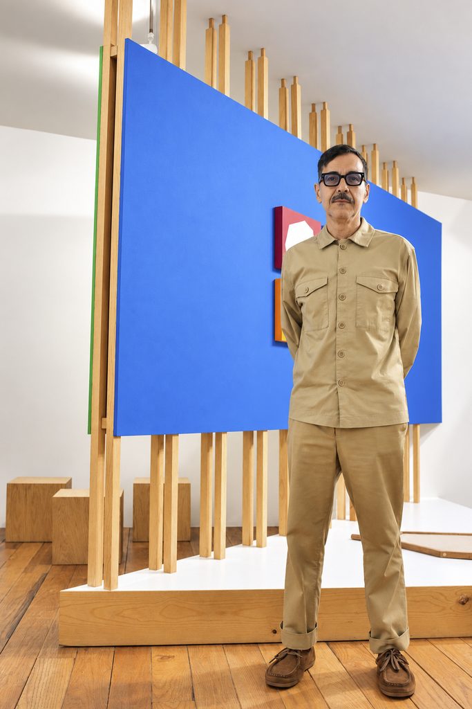 Omar Guerra M. de pie, con lentes negros y ropa beige, frente a un panel azul con pinturas geométricas y una estructura de listones de madera.