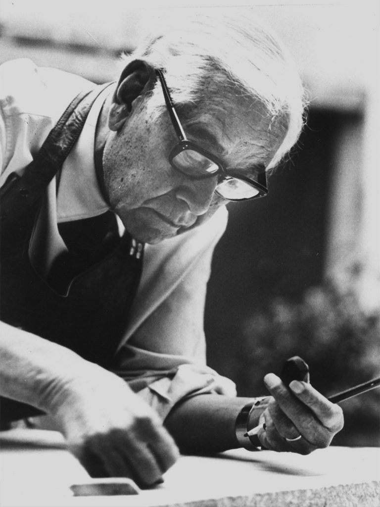 Fotografía en blanco y negro de Rufino Tamayo con lentes y delantal, inclinado sobre una superficie de trabajo mientras sostiene herramientas.