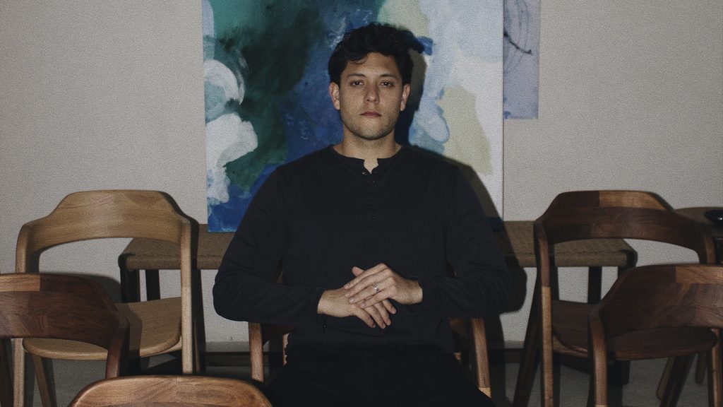 Retrato horizontal de Sebastián Ángeles sentado con las manos juntas, rodeado de sillas de madera y con una pintura abstracta al fondo.