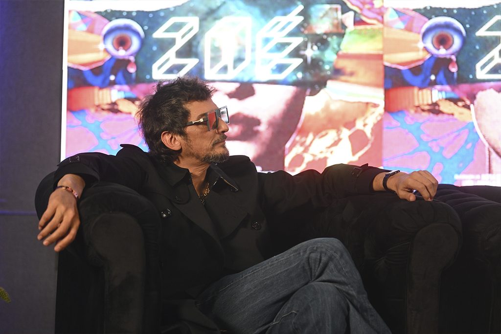 León Larregui, vocalista de Zoé, sostiene un micrófono sentado en un sillón, con una pantalla multicolor al fondo.