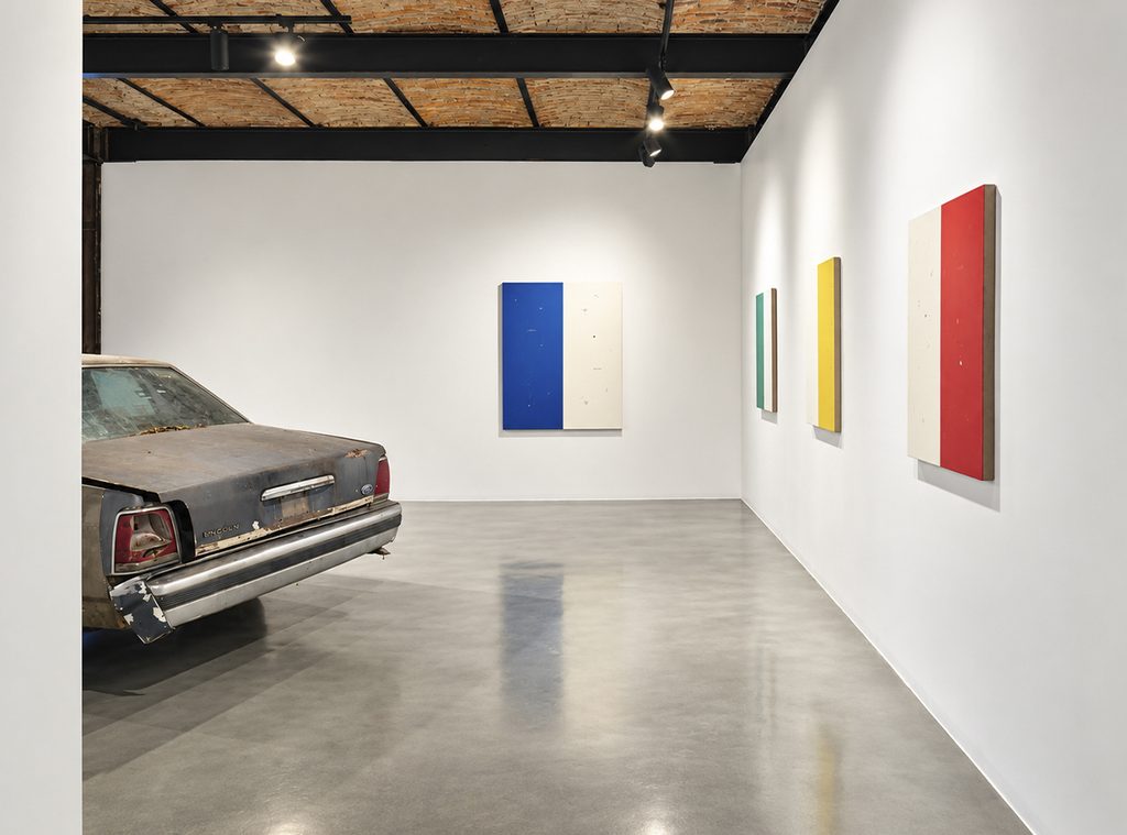 A gray car with a weathered body on display in a gallery, surrounded by paintings featuring blue, green, yellow, and red stripes.