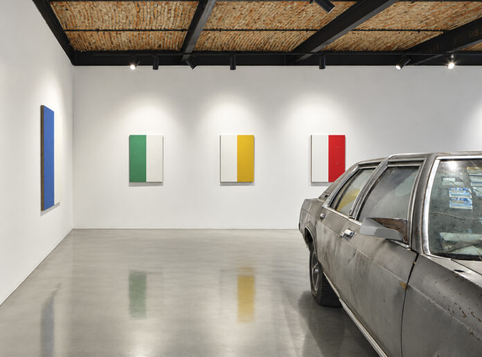 A weathered gray car next to four colorful geometric paintings in a room on Travesía Cuatro with a brick ceiling.