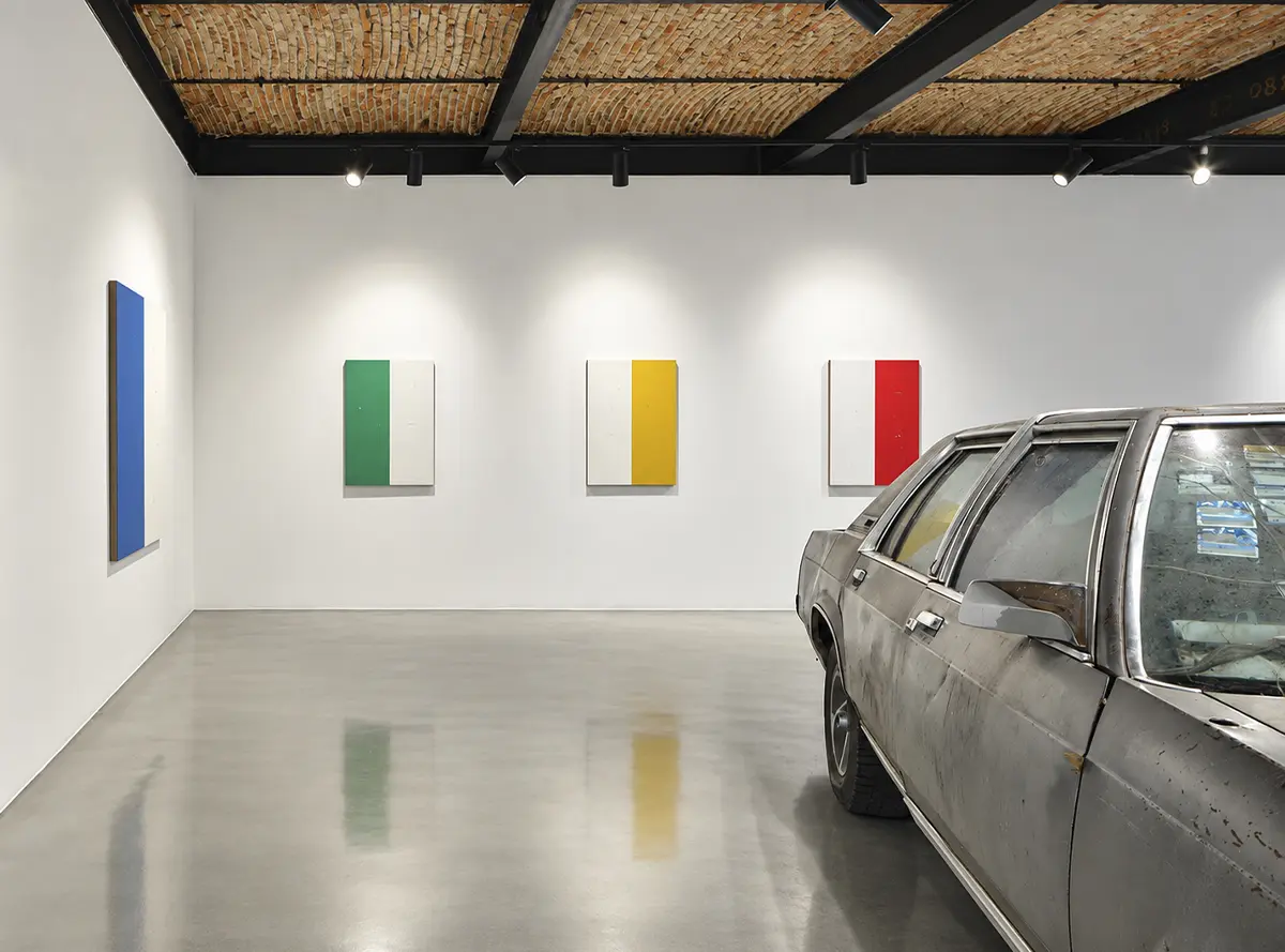 A weathered gray car next to four colorful geometric paintings in a room on Travesía Cuatro with a brick ceiling.